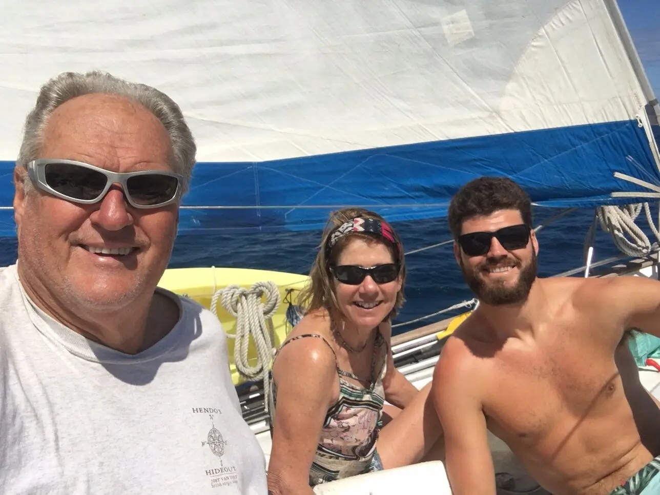 Three guests smiling on deck with the sail and deep blue ocean behind them